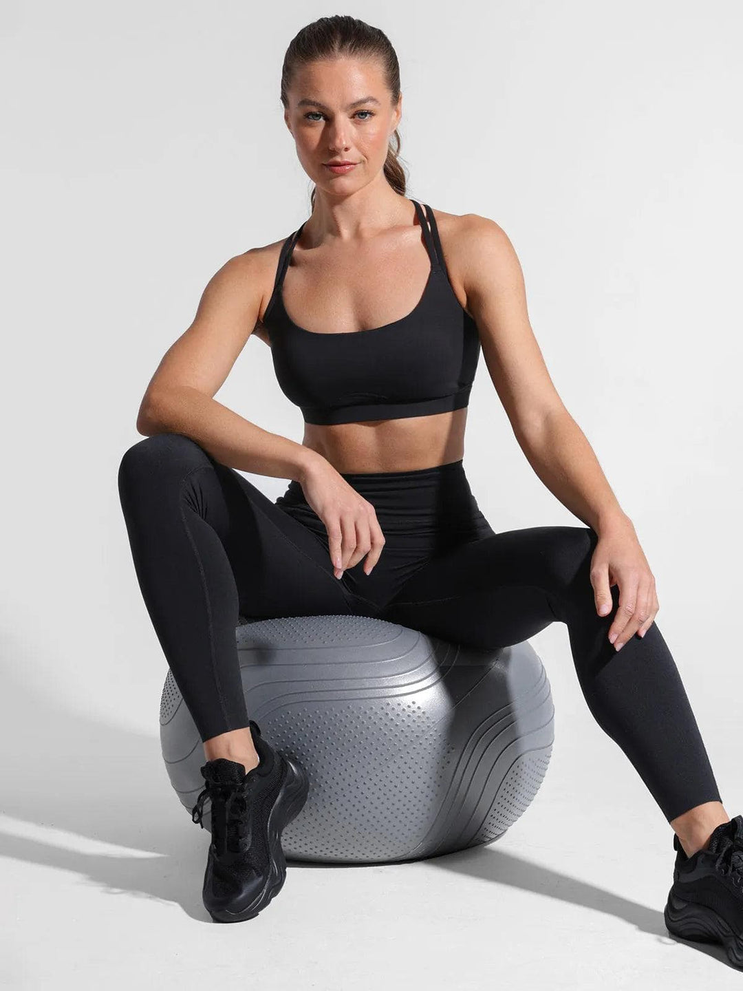 Woman in black cross-strap sports bra and high-waisted leggings sitting on a silver exercise ball against a white background, showcasing athletic fitness wear
