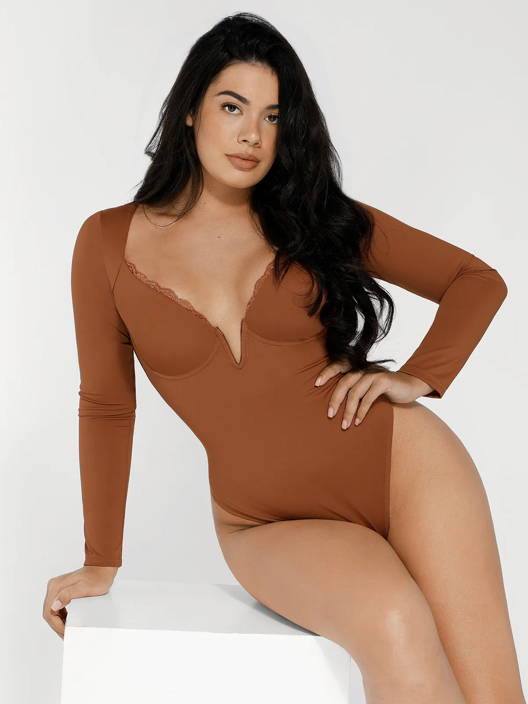 Woman wearing brown long-sleeve bodysuit with lace trim neckline, sitting on white cube against neutral background